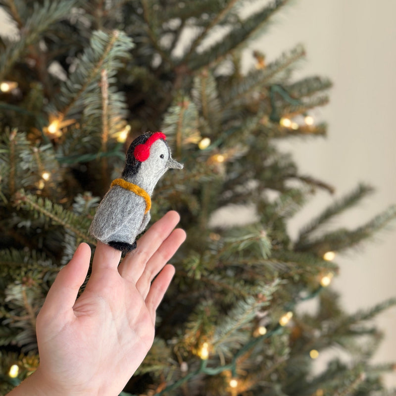 A hand holds up a handcrafted felt penguin finger puppet from a set of two, placed against a softly lit Christmas tree backdrop. This unique grey and white felt puppet for finger play is adorned with bright red headphones and a yellow scarf.