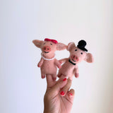 A playful set of two wool felt pig finger puppets, a charming piggy couple, are held aloft on fingers with red nail polish against a white background. The female felt pig finger puppet has a bright red bow and white pearls, while the male sports a black top hat and bow tie. Both pink pigs feature expressive stitched smiles and detailed snouts, crafted for imaginative play.