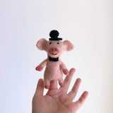 A handmade felt pig finger puppet is held by a female hand against a plain white background. This pink felt pig finger puppet, part of a piggy couple set, is adorned with a black bow tie and a black top hat. It features pink feet, ears, arms, and a snout with two nostrils, reflecting its unique design and fair-trade craftsmanship.
