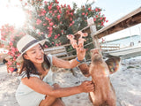 A pair of light pink felt pig finger puppets, presented as a 'Piggy Couple' set, is held up by a smiling woman in a lively outdoor beach setting. One handmade felt pig finger puppet wears a small red hat and a pearl necklace, while the other features a black hat and a bowtie. A bush with bright pink flowers in the background enhances the cheerful and fun atmosphere.