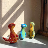 Three handmade wool felt Brontosaurus finger puppets, a playful Dino Brothers set, stand side-by-side on a sunlit windowsill, casting cozy shadows. This set includes a red and orange, a blue and green, and a green and yellow Brontosaurus, each featuring white spots, a long neck, and a small eye. Their 3D needle-felted design is showcased in the warm natural light.