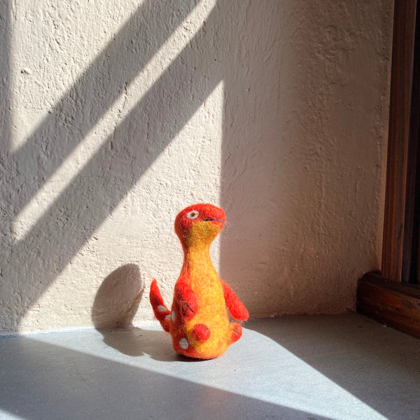 A vibrant wool felt Brontosaurus finger puppet from the 'Dino Brothers' set rests on a gray surface, bathed in natural light against a textured wall. This handcrafted needle-felted toy has a lifelike 3D design, featuring a long neck and a tail. Its body transitions from red at the head to yellow on the main body, complemented by red arms and white spots. This unique felt toy for kids is ideal for imaginative play.
