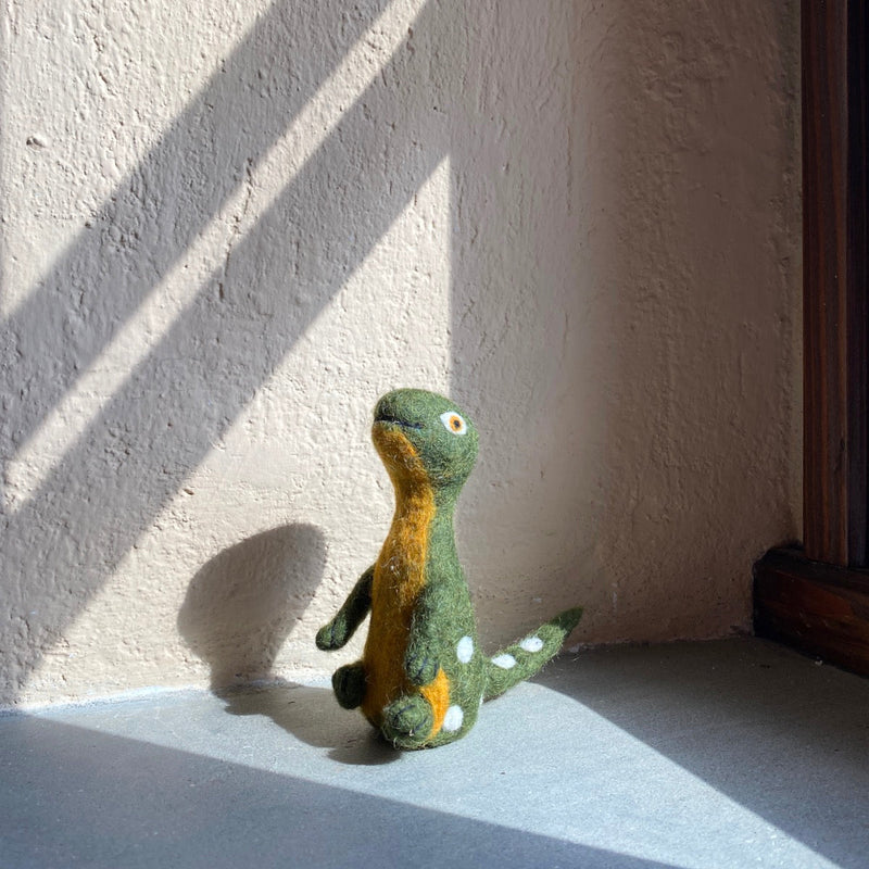 A handcrafted Brontosaurus felt finger puppet, from the 'Dino Brothers' collection, rests on a light gray surface near a wall, naturally lit by sunlight creating subtle shadows. This unique needle-felted dinosaur puppet, made from 100% wool, features a long neck, a green body with an orange underbelly, and small white dots along its back and tail. Its detailed stitching and small eye highlight its handmade, 3D design for a lifelike appearance.