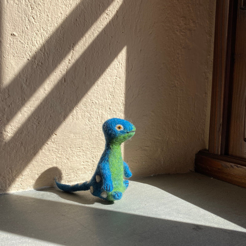 A handmade wool felt Brontosaurus finger puppet rests indoors near a wall with window shadow patterns. This unique, needle-felted dinosaur toy, part of the 'Dino Brothers' collection, features a long neck, a blue head, a green body, and white spots. Handcrafted from 100% wool felt, this puppet offers a playful and lifelike 3D design, showcasing artisanal craftsmanship.