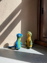 Two colorful wool felt Brontosaurus finger puppets from the 'Dino Brothers' set are showcased on a gray surface, positioned next to a brown wooden doorframe in a warm, sunlit, rustic setting. These handmade, needle felted Brontosaurus puppets, one blue and green with white spots and the other yellow and green, feature 3D, lifelike designs with long necks and tails. They stand upright, highlighting their tactile appeal and inviting imaginative play for kids.