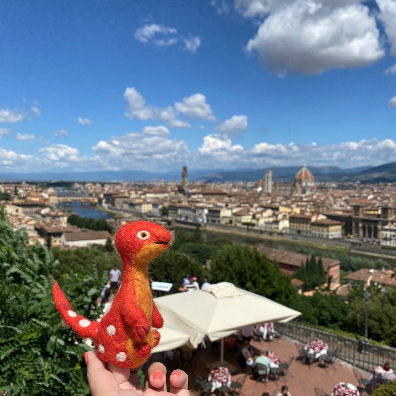 A vibrant red and yellow wool felt Brontosaurus finger puppet, part of the Dino Brothers set, is playfully held by a female hand against a bright, sunny cityscape. This unique, handcrafted felt kids toy features a long neck and small arms, with white dots detailing its tail, evoking a sense of adventure. The background shows a bustling terrace with umbrellas and city buildings under a cloudy blue sky.