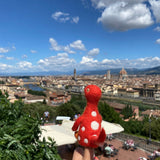 A vibrant red wool felt Brontosaurus finger puppet, part of a dinosaur finger puppet set, is held up on a finger against a bright, sunny sky and a distant, blurred cityscape. This handmade needle felted Brontosaurus features distinctive white spots, a long neck, small arms, and a tail, designed with a lifelike 3D appearance.