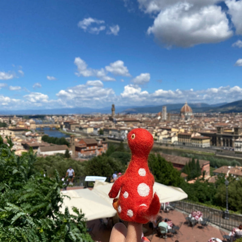A vibrant red wool felt Brontosaurus finger puppet, part of a dinosaur finger puppet set, is held up on a finger against a bright, sunny sky and a distant, blurred cityscape. This handmade needle felted Brontosaurus features distinctive white spots, a long neck, small arms, and a tail, designed with a lifelike 3D appearance.