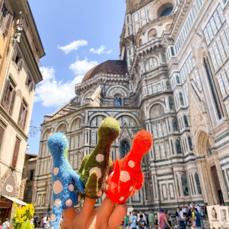 A set of three handcrafted wool felt Brontosaurus finger puppets are playfully showcased on fingers, set against a backdrop of classical urban architecture under a bright sky. These unique Brontosaurus puppets come in blue, green, and red, each adorned with white spots. Their handcrafted nature emphasizes a whimsical blend of playful toys and iconic urban scenery.
