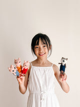 A young girl smiles, holding up a set of four handcrafted felt finger puppets against a plain, light background. On her left hand are three light pink needle felted pig finger puppets in blue, yellow, and red overalls. On her right is a gray and white wolf finger puppet in dark blue overalls. Both the playful pig and wolf puppets feature small ears and stitched facial details.