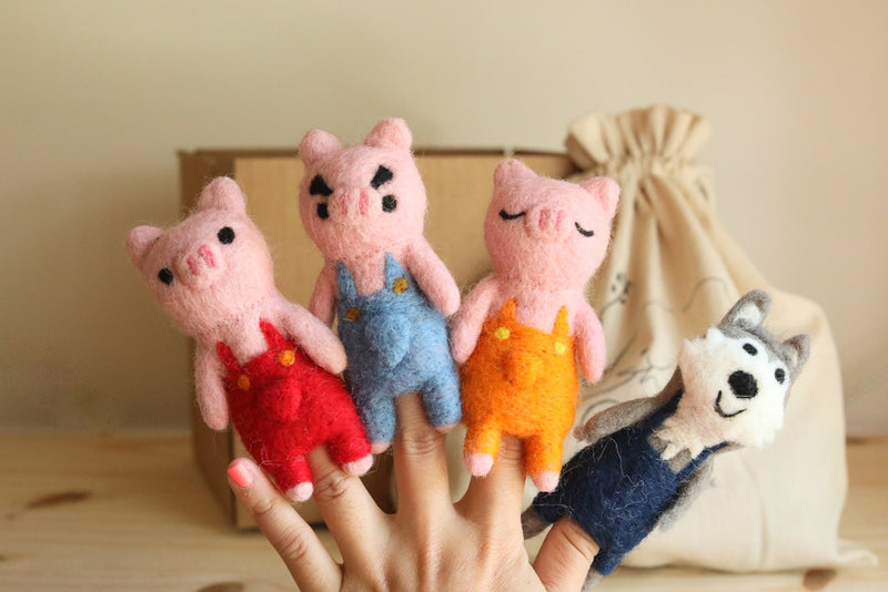 This handmade set of four wool felt finger puppets, featuring the Three Little Pigs and the Big Wolf, is playfully displayed on a hand against a neutral background. The three pink pig finger puppets have unique facial expressions and wear red, blue, and orange overalls. The gray wolf finger puppet features expressive details and dark blue overalls. These fair-trade, handcrafted felt puppets are perfect for imaginative storytelling.