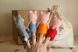 A female hand presents a set of four unique handmade felt finger puppets, against a softly blurred background. This handcrafted felt kids toy set includes three pink pig finger puppets, each wearing blue, orange, or red overalls, alongside a grey wolf finger puppet with blue overalls and a small white tail. Each wool felt finger puppet has distinct shapes and textures, evoking imaginative play.
