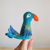 A handcrafted felt finger puppet of a parrot, resting on a human hand, features vibrant colors. The parrot is depicted in profile, facing left, with a blue body, orange beak, green head, and spread wings showcasing a feather-like pattern. Its tail extends downwards from the body, giving it a friendly expression with wide-open eyes.