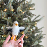 A hand holds a small, plush seagull toy made from wool felt, featuring yellow beaks and feet, large expressive eyes, and a white background. The festive setting includes a Christmas tree with lights in the background, evoking warm and cozy feelings.