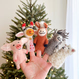 A set of five handcrafted wool felt farm animal finger puppets are playfully displayed on a hand, set against a blurred Christmas tree background with warm, glowing lights. This charming collection includes a pink pig, a white and cream cow with red horns, a brown horse with a white mane, a grey donkey, and a fluffy white sheep, all ready for imaginative play.