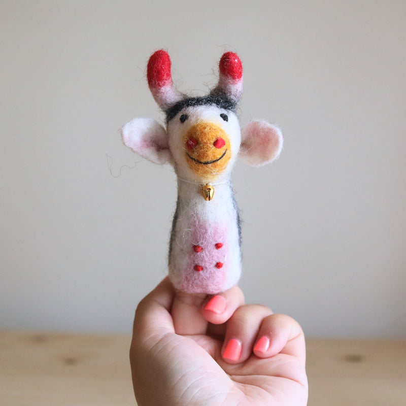 A handcrafted 100% wool felt cow finger puppet is held gently by a female hand with bright pink nails, set against a simple light beige wall and light-colored wooden surface. This soft, textured felt farm animal finger puppet features a white face, smiling orange nose, black and white hair, red-tipped horns, a gold bell, and pink spots, showcasing its unique design.