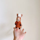 A handcrafted wool felt horse finger puppet is held upright on an open hand, presented against a plain backdrop. This charming brown horse features a white mane, black hooves, and a stitched smile, showcasing its delightful details and artisan craftsmanship.