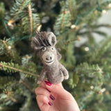 A light brown and gray handmade felt donkey finger puppet, made of wool felt, is held by a female hand with red manicured nails. The puppet features a tufted gray and brown mane and stitched black facial details. This unique kids felt toy is presented against a softly blurred Christmas pine tree with small lights, creating a playful and festive scene.