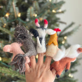 A set of five handcrafted wool felt farm animal finger puppets are displayed on a female hand, worn on her fingers, against a softly blurred Christmas tree background with warm lights. From left, the charming collection includes a light pink pig, a grey horse with a dark grey yarn mane, a white and black cow with red horns, a hen with a red comb, and a reddish-brown sheep with a white wooly fleece.