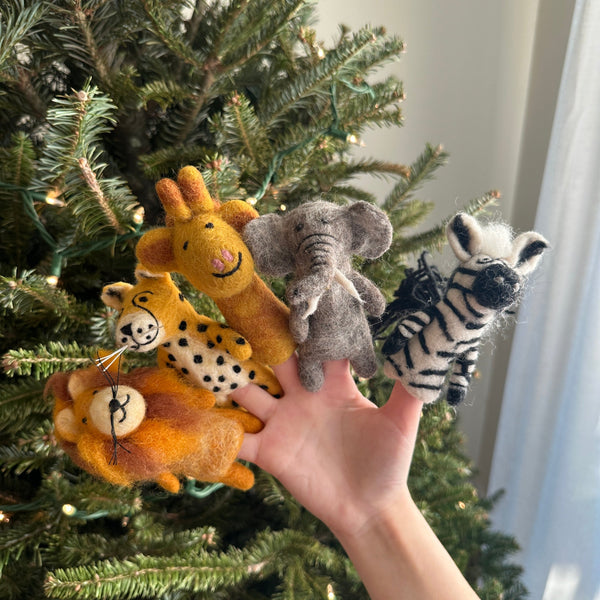 A set of five handcrafted felt safari animal finger puppets is presented by a female hand against a festive Christmas tree background. The collection features a felt lion finger puppet with a golden mane, a spotted felt cheetah finger puppet, a long-necked felt giraffe finger puppet, a gray felt elephant finger puppet, and a striped felt zebra finger puppet. These unique, fair-trade felt kids toys are ideal for imaginative play.