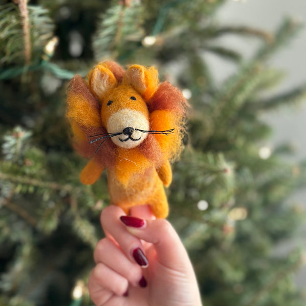 A handmade felt lion finger puppet is gently held by a hand against a blurred, festive Christmas tree with twinkling lights. This unique safari animal puppet, part of a set of five, features a fuzzy orange and brown mane, a cream muzzle with a stitched black nose, and an embroidered face. Handcrafted in Nepal, this felt toy for kids brings whimsical charm and supports fair-trade artisans.
