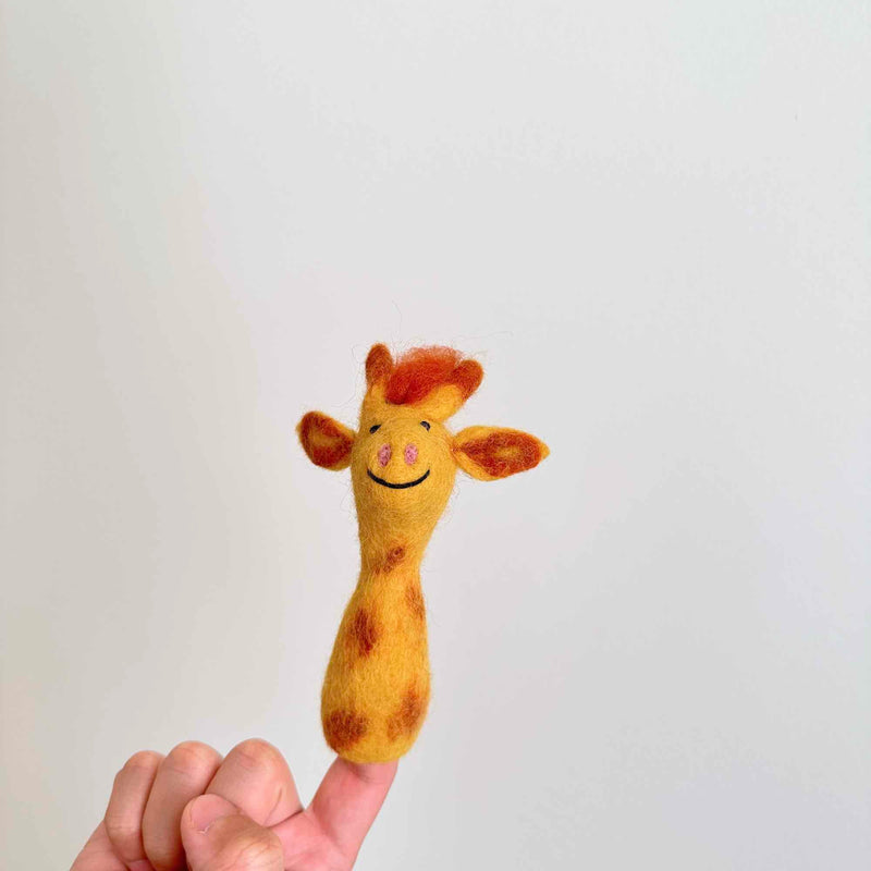 A wool felt giraffe finger puppet, from the handmade Felt Finger Puppets Safari Animals set, is playfully displayed on a human finger against a white backdrop. This charming, fair-trade giraffe puppet, 100% handmade in Nepal, features a yellow body with brown spots, small brown ears, and a tuft of brown felt hair. It has a stitched smile and pink nostrils, emphasizing its delightful and unique design.