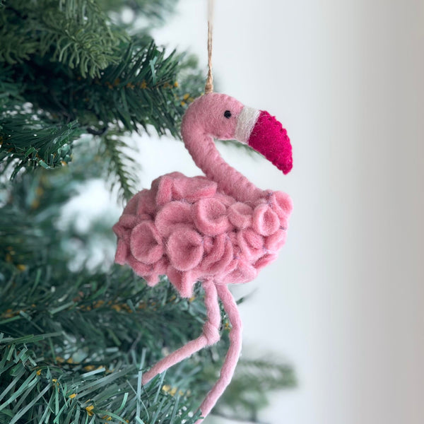A pink felt flamingo ornament, hanging from a green Christmas tree, is set against a plain white wall, evoking a festive and cheerful mood. This handmade felt flamingo ornament features a darker pink peak and a white stripe, crafted with a textured body and suspended by a jute string.