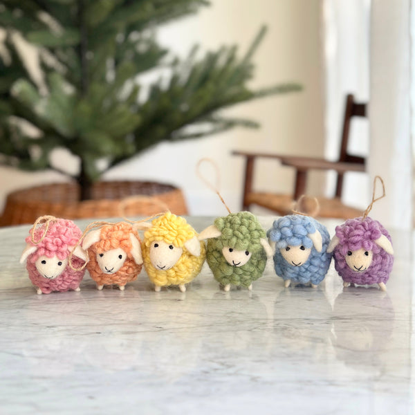 Six felt fluffy sheep ornaments, a set in pastel tones, are arranged on a marble table, with a blurred Christmas tree in the background. Each of these unique Christmas ornaments is made of felted wool, featuring a fluffy texture, small white ears, and a jute thread for hanging. The sheep come in pink, beige, yellow, green, blue, and purple, adding a playful touch to holiday decor.