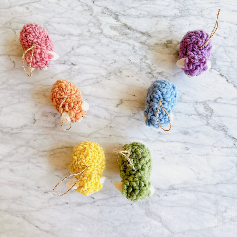 Six pastel-toned wool felt fluffy sheep ornaments are playfully arranged on a white marble backdrop. These charming handmade felt ornaments, crafted from felted wool, come in pink, beige, yellow, green, blue, and purple. Each mini sheep has a fluffy texture and a jute string for hanging, perfect for unique Christmas decor.