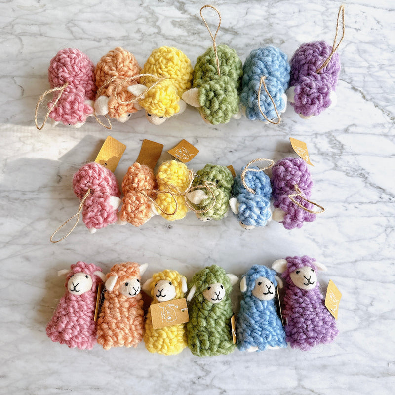 A cheerful set of six mini felt fluffy sheep ornaments is arranged in rows on a white marble surface. These soft, felted wool sheep ornaments feature fluffy textures in a variety of pastel tones, including pink, beige, yellow, green, blue, and purple. Each ornament includes a jute string for easy hanging.
