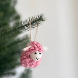A pink felt fluffy sheep ornament hangs from a dark green Christmas tree branch. This wool felt ornament has a fluffy texture, small white ears, black eyes, and a small black smile. It is suspended by a jute string, creating a calm, festive atmosphere.
