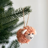 A peach felt fluffy sheep ornament hangs from a vibrant green Christmas tree branch against a clean white backdrop. This mini wool felt sheep ornament features a fluffy pastel peach body, a white face with ears, and a stitched smile. A rustic jute thread provides a charming hanger, perfect for a cozy and playful holiday display.