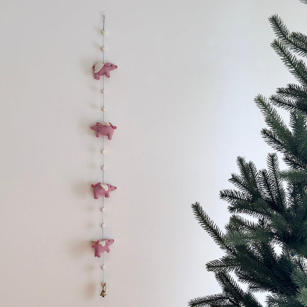 A pink felt flying pig garland is artfully displayed against a clean white wall, with a partial pine tree visible on the right, creating a whimsical scene. This playful wool felt garland features four soft pink pigs, each with small white wings, evenly spaced along a thin string, interspersed with small, round, cream-colored wool felt balls.