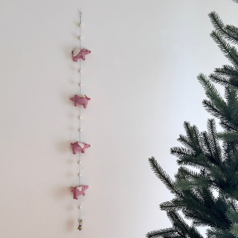 A pink felt flying pig garland is artfully displayed against a clean white wall, with a partial pine tree visible on the right, creating a whimsical scene. This playful wool felt garland features four soft pink pigs, each with small white wings, evenly spaced along a thin string, interspersed with small, round, cream-colored wool felt balls.