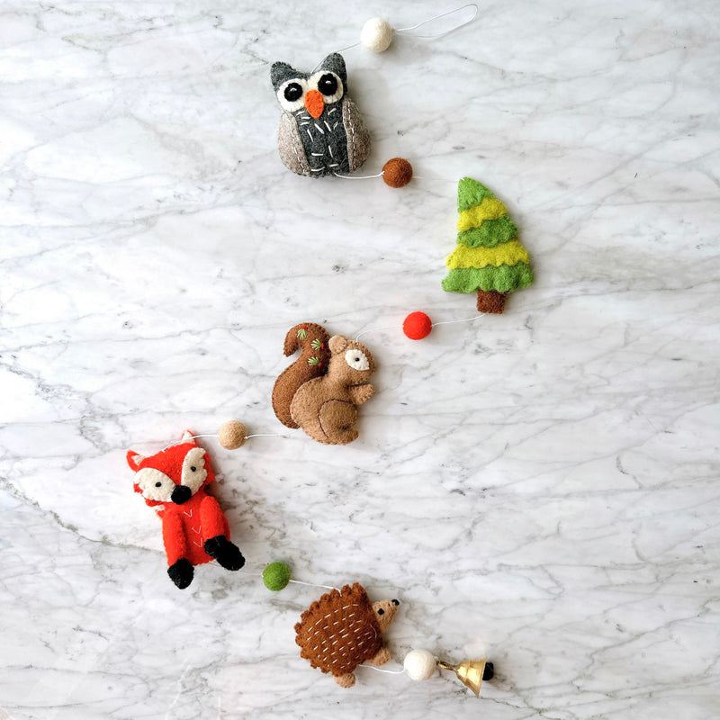 A Felt Forest Animal Garland is displayed on a marbled surface. This charming wool felt garland features detailed 3D animals including a grey and white owl, a green and brown tree, an orange fox with black feet, a brown hedgehog, and a lighter brown squirrel with red embroidery. A copper bell hangs at the end.