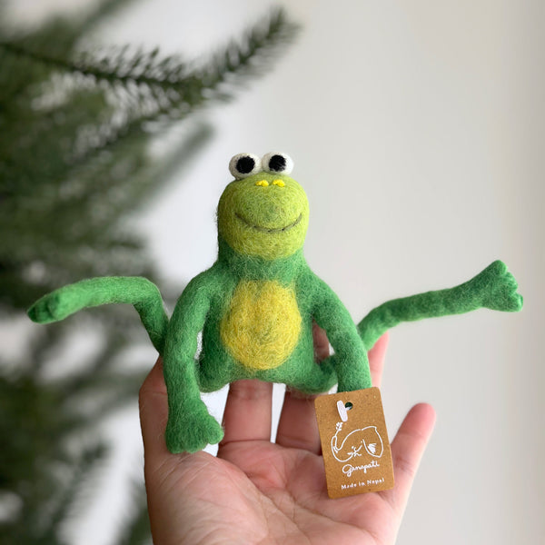 A female hand holds a green wool felt frog finger puppet against a blurred Christmas tree branch and a white wall. This handcrafted frog finger puppet is green with a yellow stomach and inner legs. It has large, expressive needle-felted eyes, a smiling mouth, and M-shaped legs. A 'Made in Nepal' tag underscores its fair-trade craftsmanship.