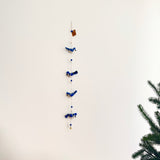 A blue jay wool felt garland hangs vertically on a plain white wall. It features four rich blue blue jay figures, each with fluffy fur on their heads and positioned in varying poses. The figures are strung along with spherical beads, topped by a small tag. To the right, green pine branches enhance the natural setting.