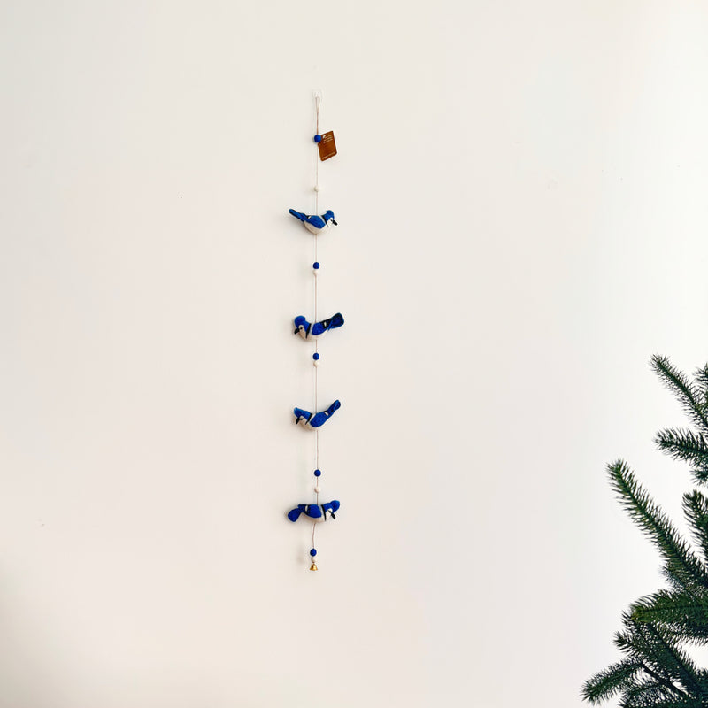 A blue jay wool felt garland hangs vertically on a plain white wall. It features four rich blue blue jay figures, each with fluffy fur on their heads and positioned in varying poses. The figures are strung along with spherical beads, topped by a small tag. To the right, green pine branches enhance the natural setting.
