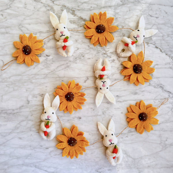 A felt Bunny and Sunflower garland is laid out against a textured marble backdrop. The garland features alternating sunflowers with multiple orange petals and brown centers, and white bunnies holding a carrot, all connected by a thin jute rope.
