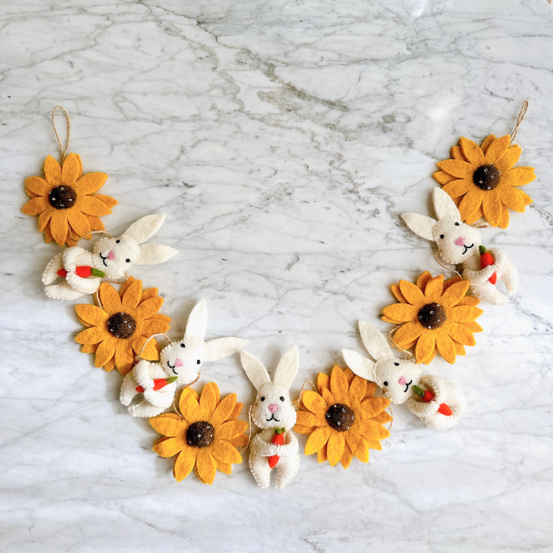 A cheerful felt Bunny and Sunflower garland is displayed in a semi-circular shape on a white marble surface. This 36-inch long felt garland features 6 alternating bright yellow sunflowers with dark brown centers and 6 white bunnies. Each white bunny playfully holds a small orange carrot, adding a delightful touch.
