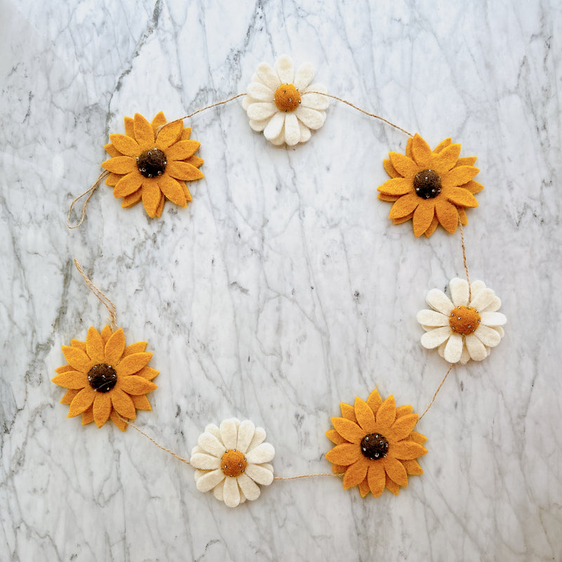 A felt Daisy & Sunflower garland is gently curved on a mottled white and gray marble background. It features alternating bright yellow sunflowers with dark brown centers and white daisies with yellow centers, all strung on a thin jute string.