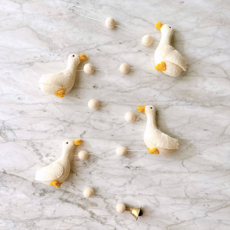 A cheerful felt duck garland is playfully arranged on a white marble surface. This handmade garland features four evenly spaced, soft off-white felt ducks with yellow beaks and feet. White felt balls are uniformly strung between the ducks on a delicate white string, with a small copper bell at one end and a hanging loop at the other.