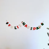 A Feliz Navidad felt garland hangs against a plain white wall, attached by clear hooks. The festive wool felt garland spells out 'Feliz Navidad' in plump red, green, and white letters, connected by jute twine. Each end is adorned with felt holly leaves and red berries, creating a celebratory display.
