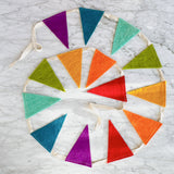 A vibrant felt Flag Party Banner garland is artfully arranged in a spiral on a white marble surface, creating a playful and festive mood. The garland features numerous triangular felt flags in shades of purple, teal, mint green, lime green, orange, yellow, and red, all attached to an off-white woven string. Each felt flag has a distinct, inviting texture.