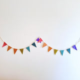 A vibrant felt flag garland, serving as a party banner, is strung horizontally on a plain white wall. This cheerful felt banner features a string of solid-colored flags in bright red, orange, yellow, teal, and purple, perfect for a festive celebration.
