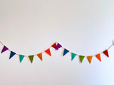 A vibrant felt flag garland, designed as a festive party banner, hangs against a bright white wall. This cheerful banner features a series of simple triangular felt flags in a variety of colors including purple, blue, green, orange, and red, strung along a neutral cord. Its playful design evokes a sense of celebration.