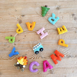 A playful felt Happy Camper garland lies flat on a wooden table. It spells out 'HAPPY CAMPER' in colorful felt letters of pink, orange, yellow, green, and blue. The garland also includes distinct felt shapes: a blue minivan, a green tree, and a red and yellow campfire. This delightful design evokes a sense of outdoor adventure.