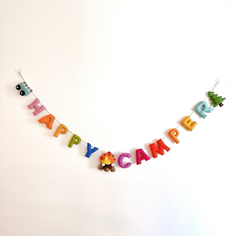 A 'Happy Camper' felt garland is displayed against a plain white wall. The garland spells 'Happy Camper' in vibrant pink, orange, yellow, green, and blue letters. It features a blue mini van on the left, a campfire in the center, and a green tree on the right, all strung on thin twine.