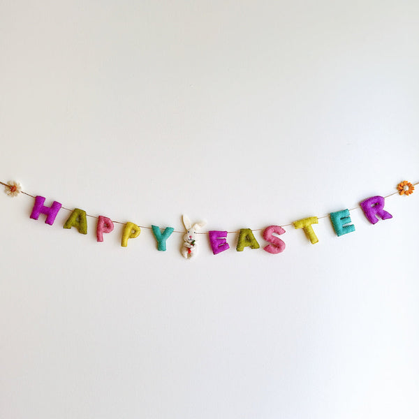 A vibrant wool felt Happy Easter garland hangs against a plain white wall, creating a cheerful and festive mood. It features individual pastel wool felt letters spelling 'Happy Easter' strung on a thin jute thread. Each end is adorned with a small wool felt daisy and sunflower. A white wool felt bunny clutching a tiny orange carrot is attached between the words, adding a playful touch.
