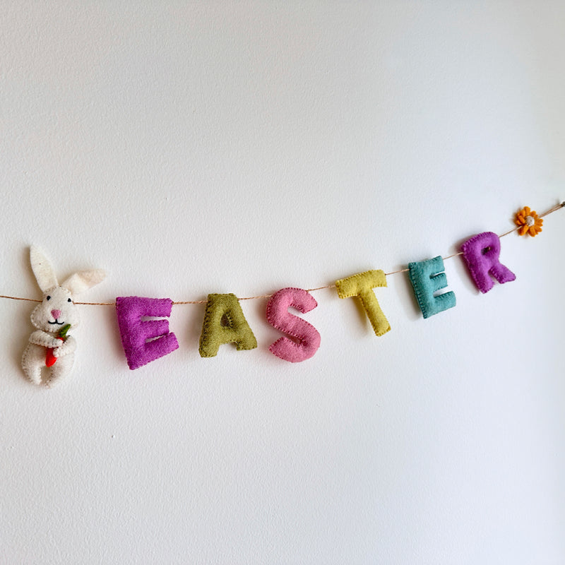 A pastel tone 'Happy Easter' wool felt garland is displayed against a clean white wall. The handmade garland spells out 'Happy Easter,' with each letter in a whimsical pastel color like purple, green, pink, yellow, and teal. It is strung on a jute string and adorned with a white felt bunny holding a felt carrot, a small orange felt sunflower, and felt daisies, adding a playful springtime charm.